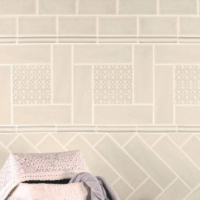 Subway tiles from the Adex Ocean Ceramic Tile collection in a soft neutral hue, featuring decorative insets with geometric patterns, accompanied by a plush folded towel and floral accents, creating a peaceful and elegant bathroom ambiance.