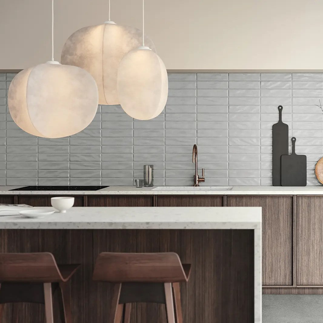 Modern kitchen interior with muted grey ceramic tiles from Surface Group, featuring oversized pendant lights and sleek wooden bar stools, showcasing a blend of warm ambiance and chic design.