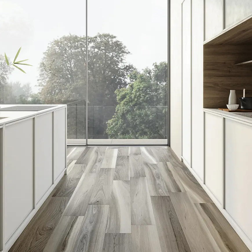 A modern interior with wood-look porcelain tiles in mixed grey and brown tones laid in a staggered pattern, complementing the sleek white cabinetry and natural wooden wall, against a backdrop of large glass windows overlooking a tranquil outdoor setting.