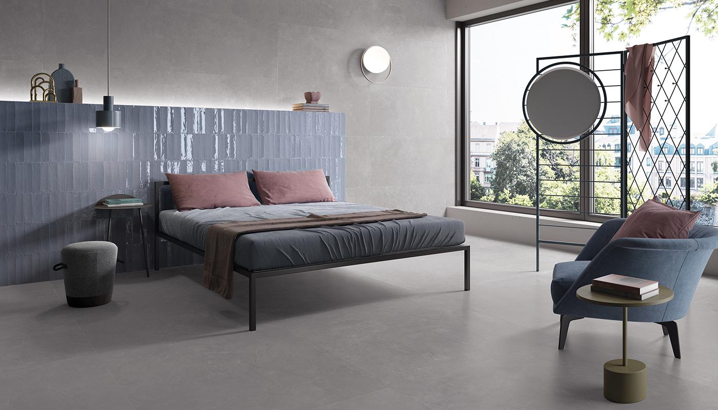 Modern minimalist bedroom interior with Italian porcelain tile flooring and wall, featuring a stylish bed, blue accent chair, and decorative elements.