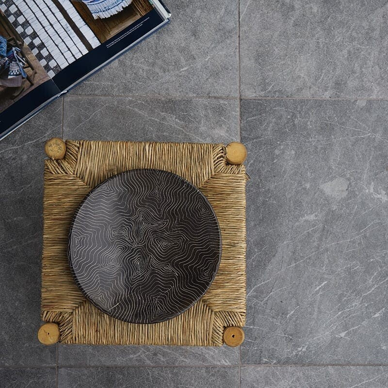 Top view of a gray tile floor with a woven stool and decorative plate. Open book with tile images in corner.