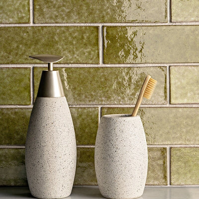 Close-up of a bathroom scene with green brick-style tile backsplash. Two beige ceramic accessories: soap dispenser and toothbrush holder with bamboo toothbrush.