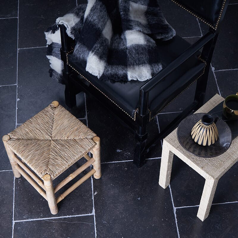 Dark room with square field tiles, woven stool, black chair with checkered blanket, beige side table with decor.