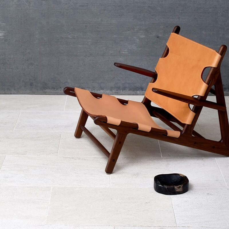 Modern room with beige marble tiles in a staggered pattern, dark gray textured wall, mid-century wooden chair, tan leather straps, small black bowl.