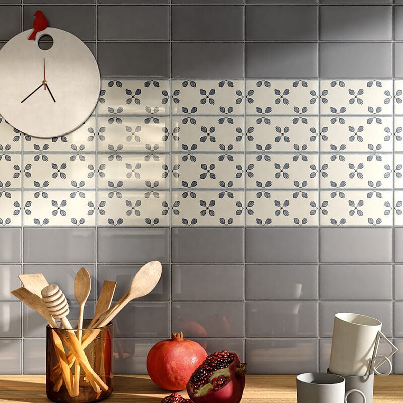 Grey rectangular tiles with beveled edge cover the wall, white tiles with blue floral strip. Modern round wall clock, glass container with wooden utensils, pomegranate, white cups.