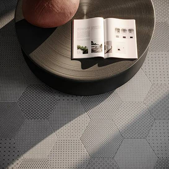 A top-down view of hexagonal porcelain tiles in shades of grey creating a honeycomb pattern on the floor, with a part of a round black and gold table visible on the left side, and an open magazine resting on the table.