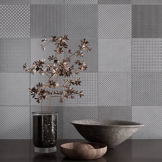 Gray silk-look porcelain tiles with a patterned design on a wall, featuring a decorative metallic branch, a black vase with a dotted pattern, and a shallow wooden bowl on a flat surface in the foreground.